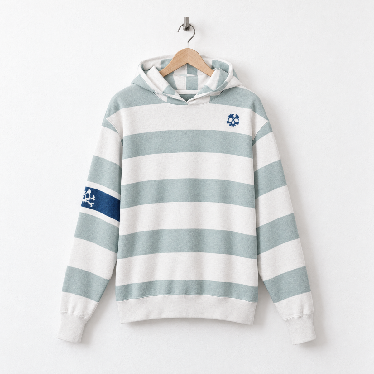 A long-sleeve hooded sweatshirt with horizontal sage green and white stripes, featuring a small blue skull-and-crossbones emblem on the left chest and a matching sleeve patch, hanging on a wooden hanger against a white wall.