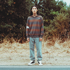 A person stands outdoors on a road with dry grass and trees in the background, wearing a brown and gray striped long-sleeve shirt, light blue jeans, and black-and-white sneakers.