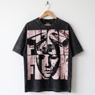 A black acid-wash graphic t-shirt hanging on a wooden hanger against a plain white wall. The shirt features a large, distressed graphic print of a stylized face and the word 'FASHION' in a pink and white block pattern.