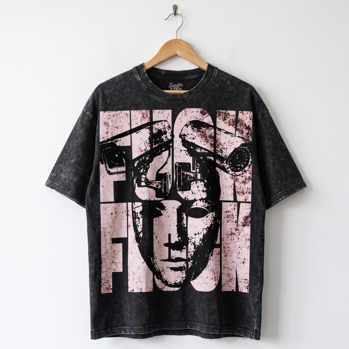 A black acid-wash graphic t-shirt hanging on a wooden hanger against a plain white wall. The shirt features a large, distressed graphic print of a stylized face and the word 'FASHION' in a pink and white block pattern.