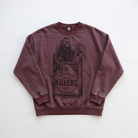 A washed dark brown crewneck sweatshirt featuring a large graphic print of a skeleton in a cigarette pack labeled 'KILLERS' on the front, laid flat on a white background.